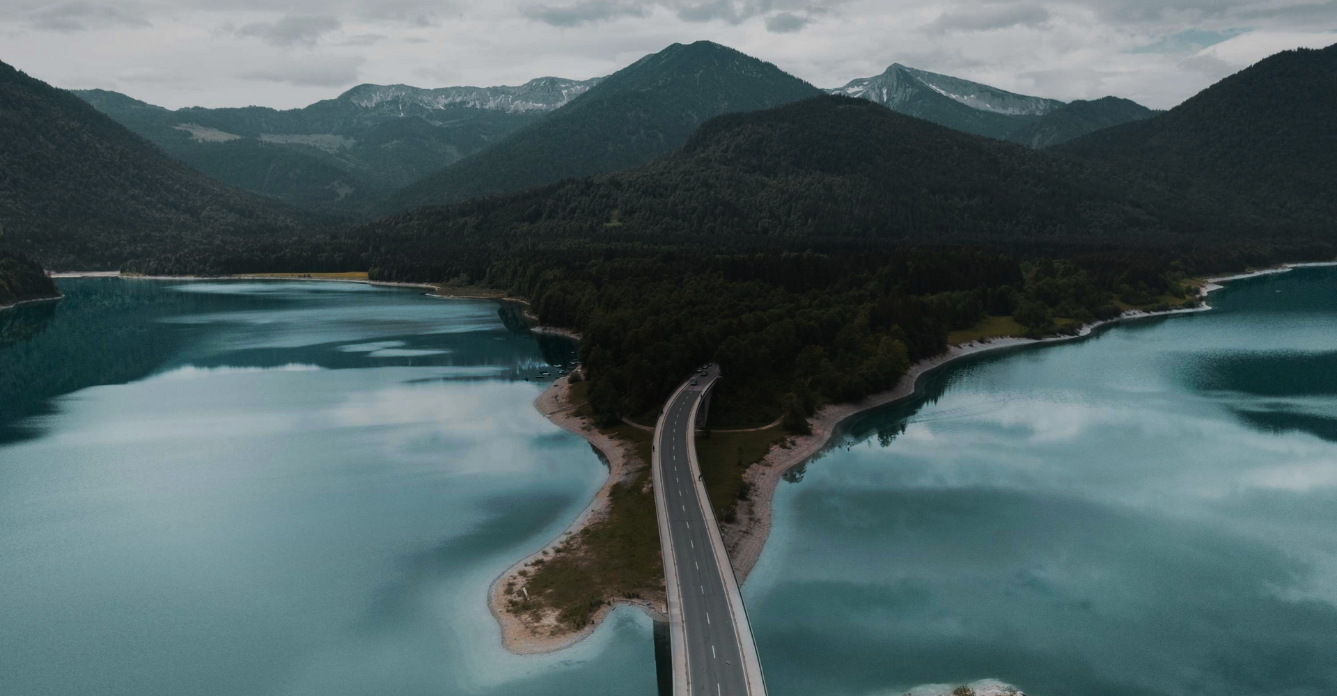 Aerial view of a narrow road leading through a lush green landscape with mountains in the background. The road is bordered by a vibrant blue lake on both sides, creating a picturesque and serene scene. The sky is overcast, adding a calm atmosphere.