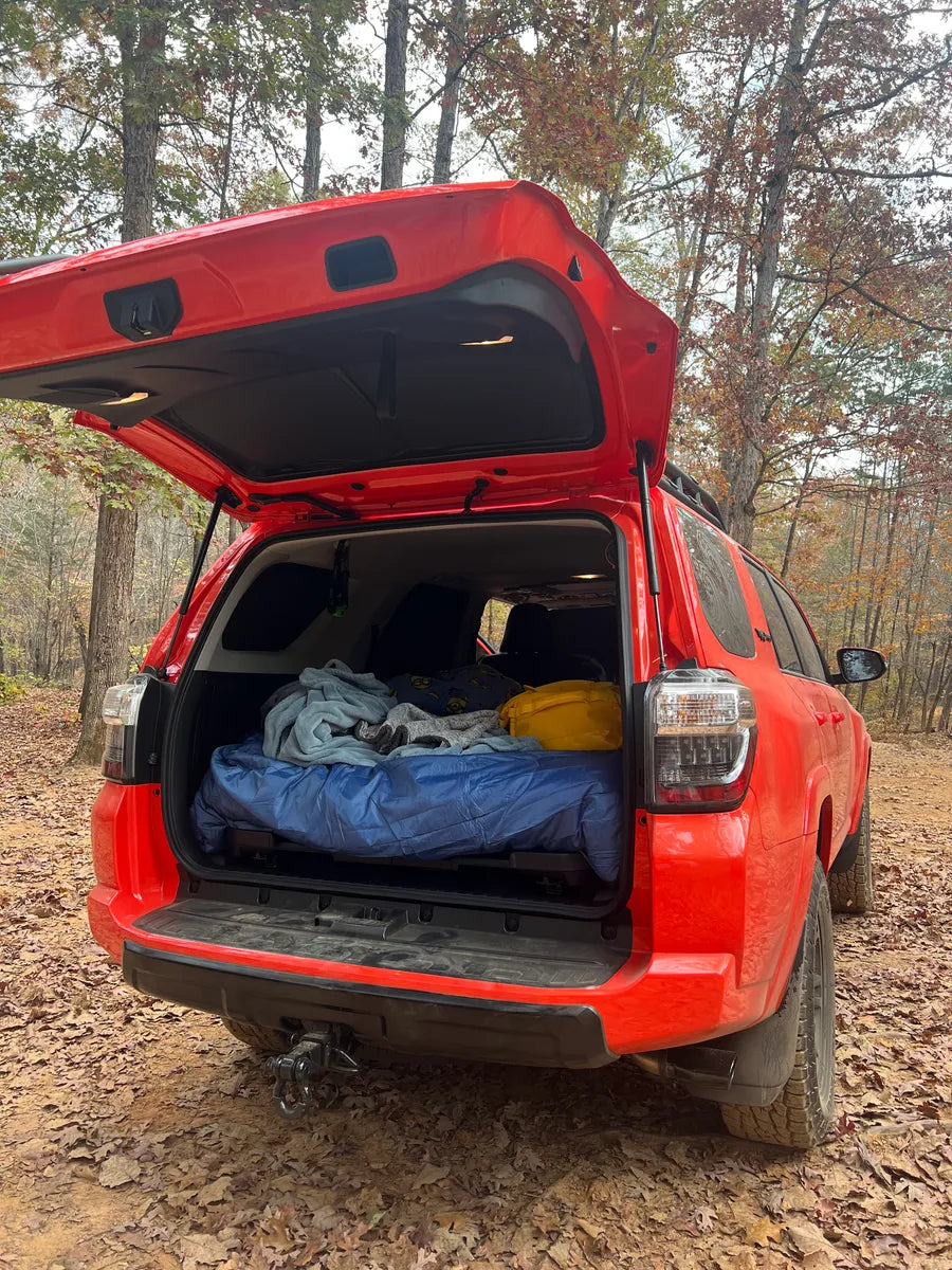 A red SUV with its trunk open, showing a neatly made bed inside, prepared for camping. The vehicle is parked in a wooded area, surrounded by trees with autumn foliage.
