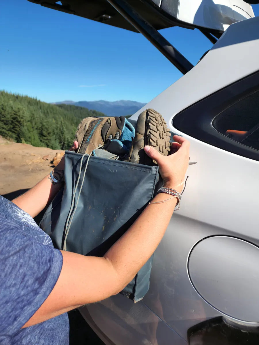 Person in a blue shirt places hiking boots into a bag attached to the side of a white vehicle in a scenic outdoor setting with trees and mountains in the background.