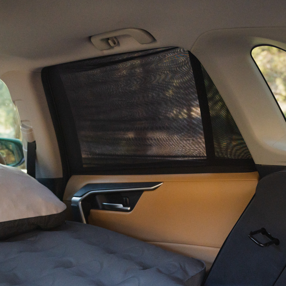 View of the interior of a car, focusing on the backseat with a sunshade attached to the window. There's a pillow and a mattress on the seat, suggesting a setup for resting or sleeping. The door's armrest and handle are visible, and trees are seen outside the window.