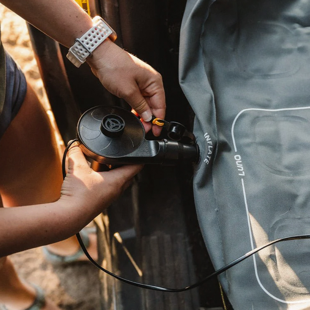 A person uses a black, portable air pump to inflate a gray air bed or inflatable mattress. The person's hands are focused on attaching the pump to the inflation valve. They are wearing a white, perforated wristwatch and light blue slippers. Sunlight brightens the scene.