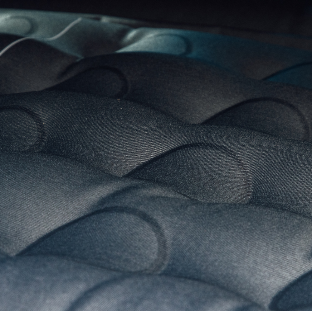 Close-up image of a dark, padded fabric surface with a quilted, bubble-like texture. The fabric has a soft, velvety appearance with gentle lighting creating gradient shadows across the curved shapes. The color appears to be a mix of dark blue and gray.