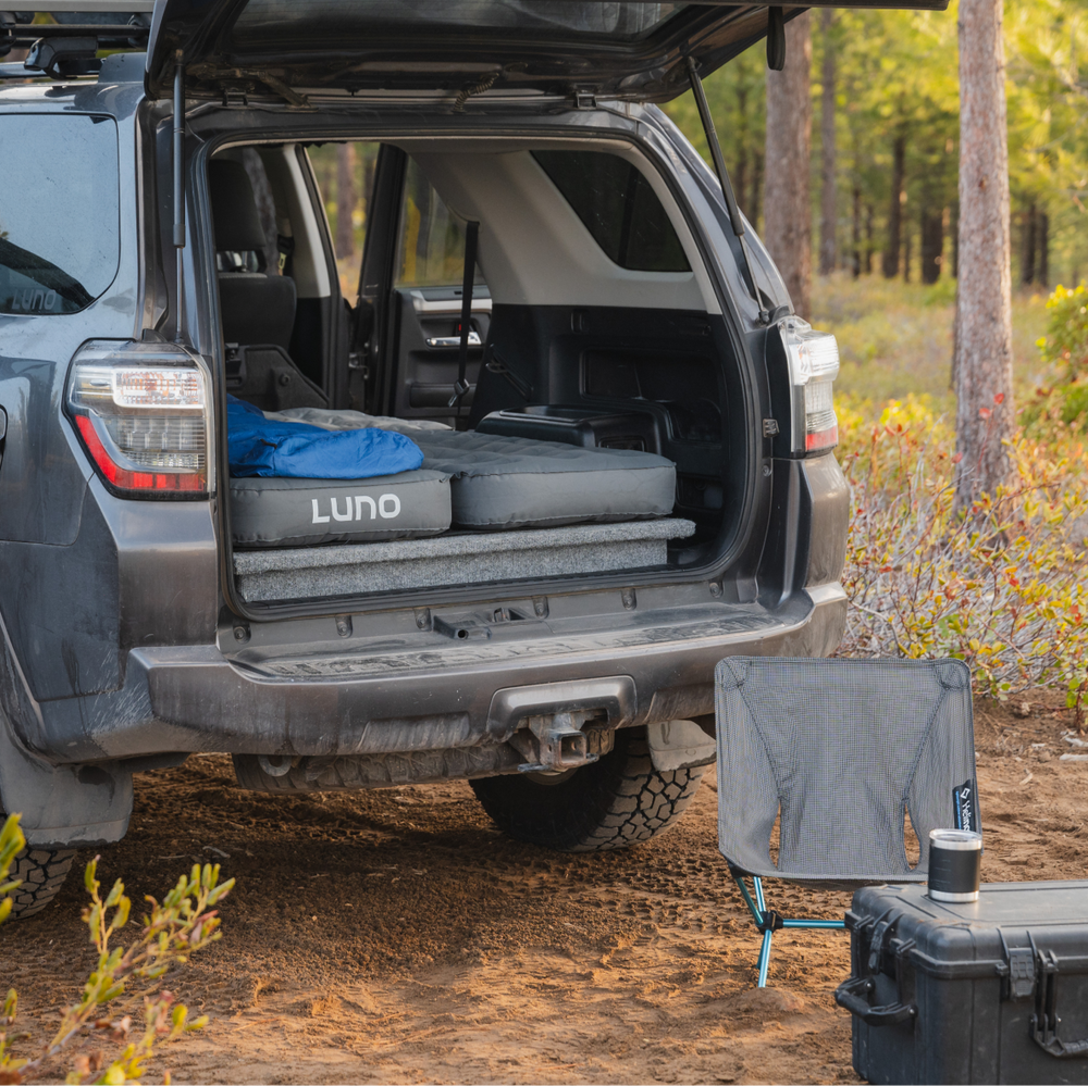 A parked SUV with the rear hatch open reveals a folded Luno mattress inside. Outside the SUV, there is a folded gray camping chair set up on the ground beside a black storage case. The background features a forest with trees and underbrush.