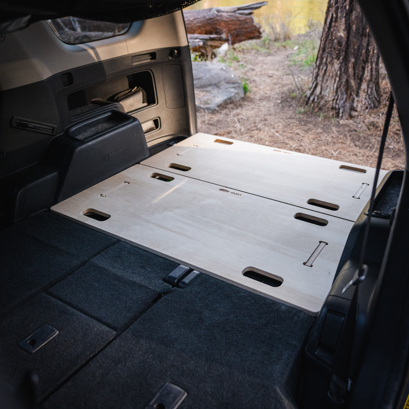 The image shows the interior of a vehicle, likely equipped with a BamBeds Vehicle Sleeping Platform. The back seats are folded down, revealing a flat, wooden platform that covers the cargo area. This platform features cut-out handles and seams, indicating that it can be adjusted or folded. In the background, trees and foliage can be seen outdoors.