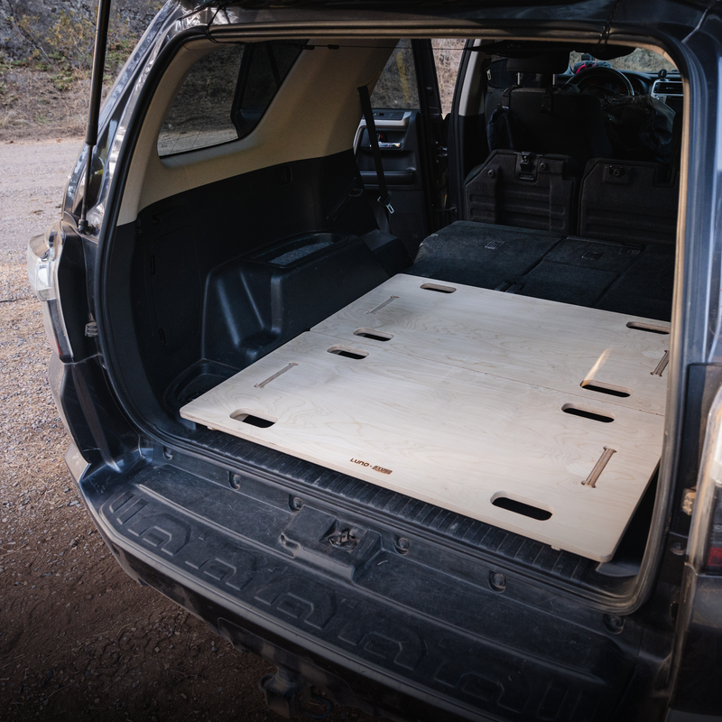 The image features the rear section of an SUV, possibly a Toyota 4Runner, with the rear door open. Inside the cargo area, a BamBeds Vehicle Sleeping Platform is custom-fitted and laid flat, covering the floor space with multiple slots and cutouts. The seats are folded down, indicating a BamBeds collaboration for optimal space utilization.