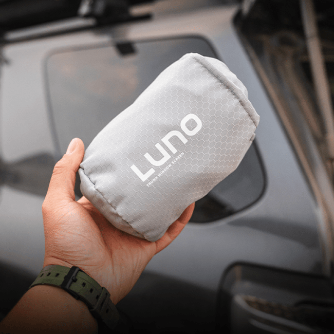 A person holds a compact, gray pouch with the "Luno HQ" logo printed on it, making it an ideal essential for car camping. In the background is a blurred view of a dark-colored vehicle, which could be perfect for using the 4Runner Trunk Window Screen (Pre-Owned).