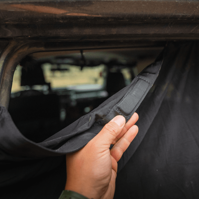 A close-up of a hand pulling back a black fabric curtain inside a vehicle unveils the interior through a window. The Luno HQ 4Runner Trunk Window Screen (Pre-Owned) is secured at the top with Velcro, serving as an essential car camping accessory with its mosquito-proof mesh. The blurred vehicle interior forms the background.