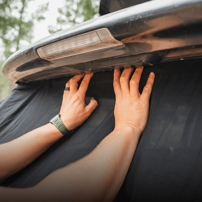 A person's hands reach under the lifted hatchback of a 4Runner, touching the underside. Sporting a green watch, they are likely setting up a 4Runner Trunk Window Screen by Luno HQ—an essential for car camping. Trees blur in the background, hinting at an adventure ready to unfold.