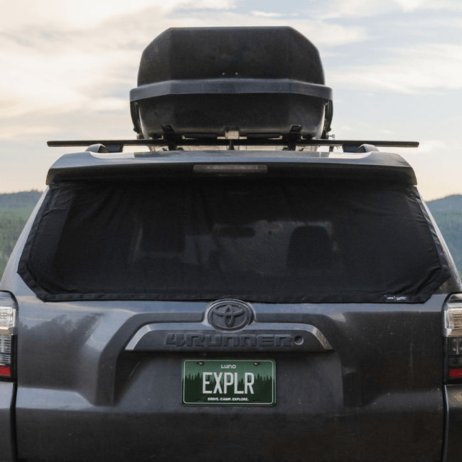 A dark gray Toyota is seen from the rear, equipped with a rooftop cargo box and a Luno HQ 4Runner Trunk Window Screen (Pre-Owned), emphasizing its car camping essentials. The license plate displays "EXPLR," and a black cover conceals the rear window. The backdrop includes a partially cloudy sky and green hills.