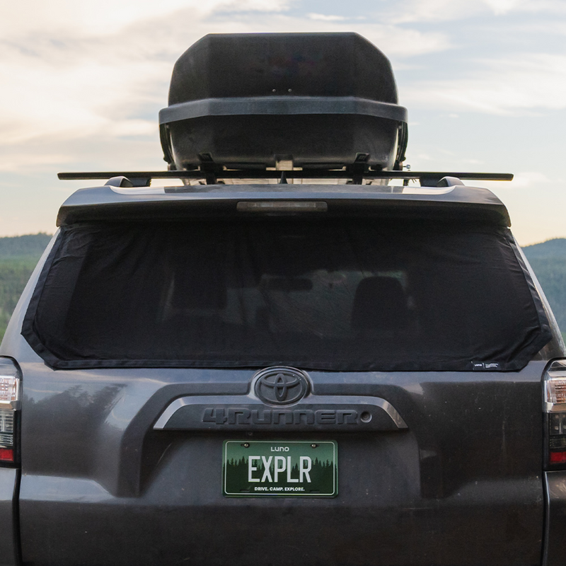 Rear view of a dark-colored SUV with a roof cargo box and a Luno®-branded license plate that reads "EXPLR." The SUV, equipped with the car camping essential Luno® 4Runner Trunk Window Screen, is parked in front of a scenic outdoor backdrop with a partially cloudy sky and distant hills.