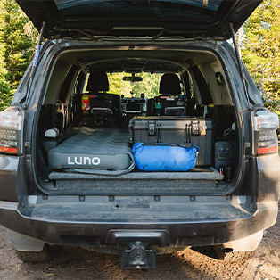 A car with its trunk open displays camping gear, including an inflatable mattress labeled "LUNO," a blue duffle bag, and other various equipment. The car is parked outdoors, surrounded by greenery.