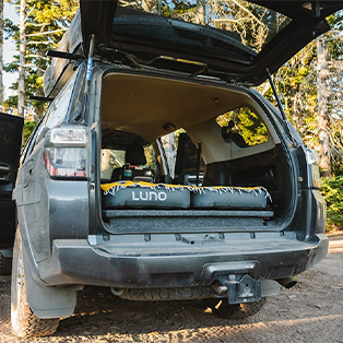 A grey SUV is parked with its rear door open, revealing a Lund-branded air mattress laid out in the back. The vehicle is surrounded by trees, suggesting a forest or wooded area.