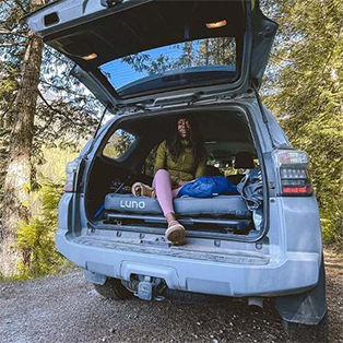 A person sits in the back of an open SUV parked in a wooded area. The SUV is equipped with a bed setup, blankets, and pillows. Trees and foliage surround the vehicle, and the person appears relaxed, enjoying the outdoor setting.