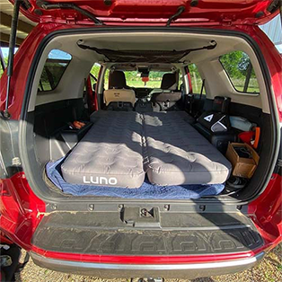 A red SUV with its back hatch open, showing an inflated air mattress labeled "LUNO" filling the cargo area. The back seats are folded down to accommodate the mattress, and camping gear can be seen around the edges. The scene is outdoors with trees in the background.