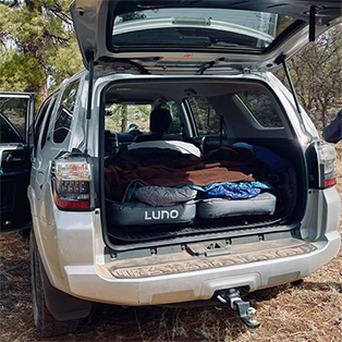 A white SUV with its rear hatch open, displaying a neatly arranged sleeping area equipped with a Luno air mattress and bedding. The vehicle is parked in a wooded area, suggesting a camping setup.