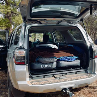 A silver SUV with its rear hatch open reveals the interior turned into a sleeping area with a Luno air mattress spread out. The vehicle is parked outdoors among trees, suggesting a camping trip. Various blankets and pillows are on the mattress.