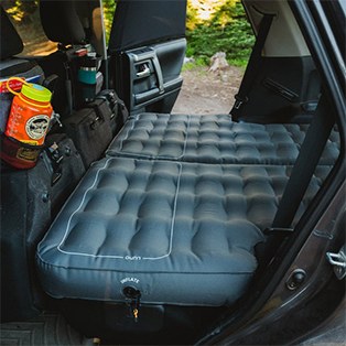 A gray inflatable mattress is set up on the back seats of a car, creating a flat and cushioned sleeping area. The rear door is open, showcasing outdoor surroundings with trees. Camping gear, including water bottles, is visible on the car's interior.
