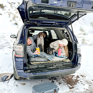 Two people and two dogs are relaxing in the open trunk of a blue SUV parked in a snowy area. They are bundled up in warm clothing, holding drinks, and appear to be enjoying the winter scenery. The hatchback of the SUV is open, revealing a cozy setup inside.