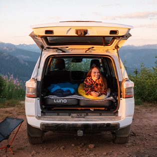 A person with long hair sits smiling in the back of a white SUV, which is parked with its tailgate open in a scenic mountainous area. Inside the SUV, there is a mattress and sleeping gear. The sun is setting, casting a warm glow. There's a camp chair nearby.