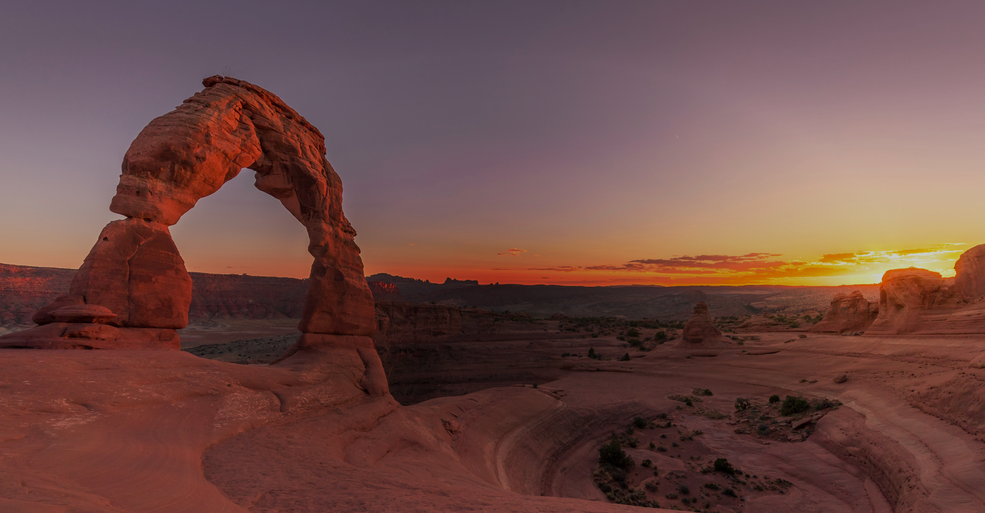 A stunning sunset view of Delicate Arch in Arches National Park, Utah. The sky takes on hues of orange and purple as the sun sets, casting shadows over the rocky landscape and emphasizing the dramatic, freestanding natural arch.