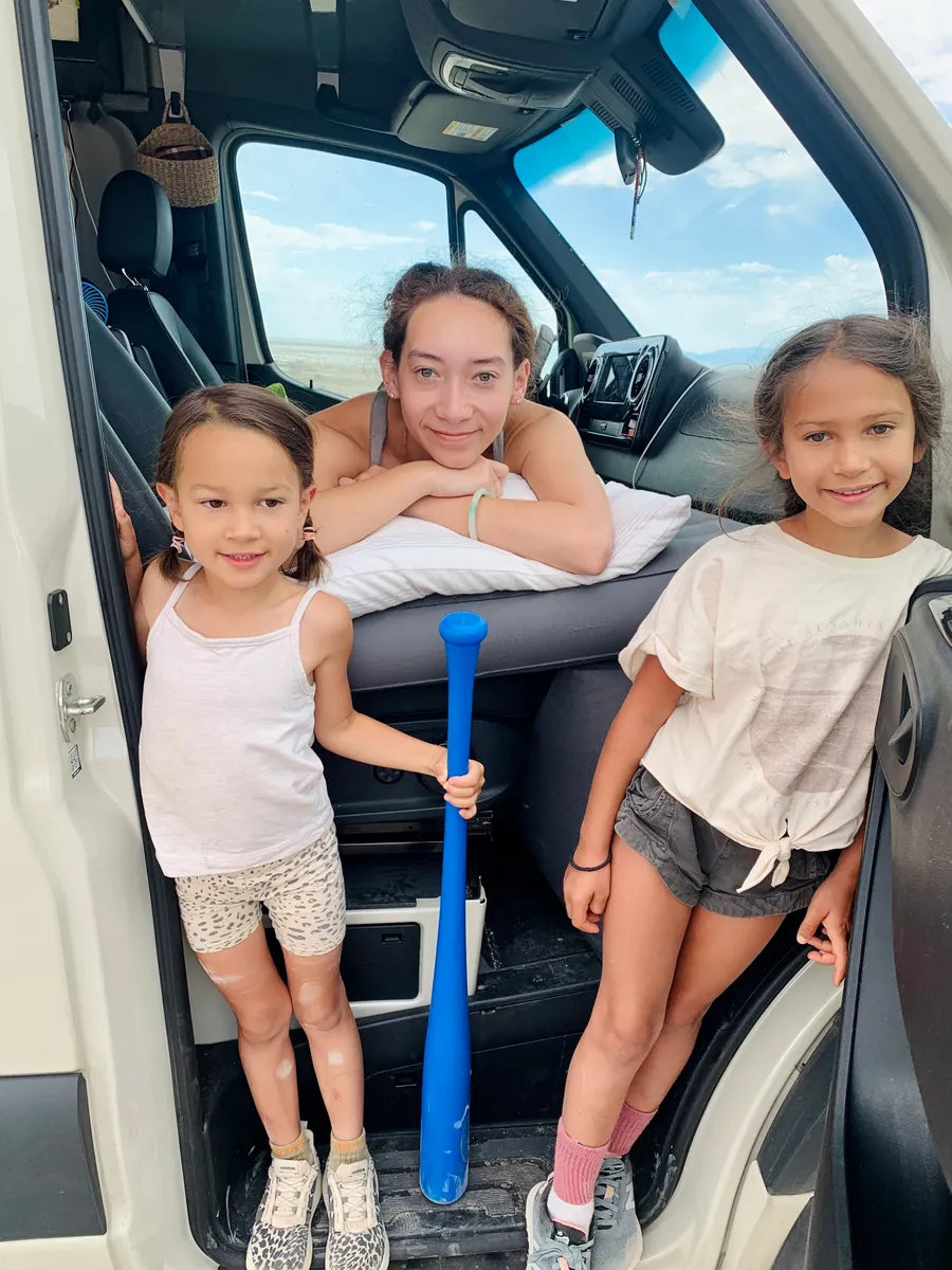 A woman leans on a van's dashboard, smiling at the camera. Two young girls stand in the doorway, one holding a blue plastic bat. Both girls wear casual summer clothing and look at the camera with cheerful expressions. The background shows a blue sky with clouds.