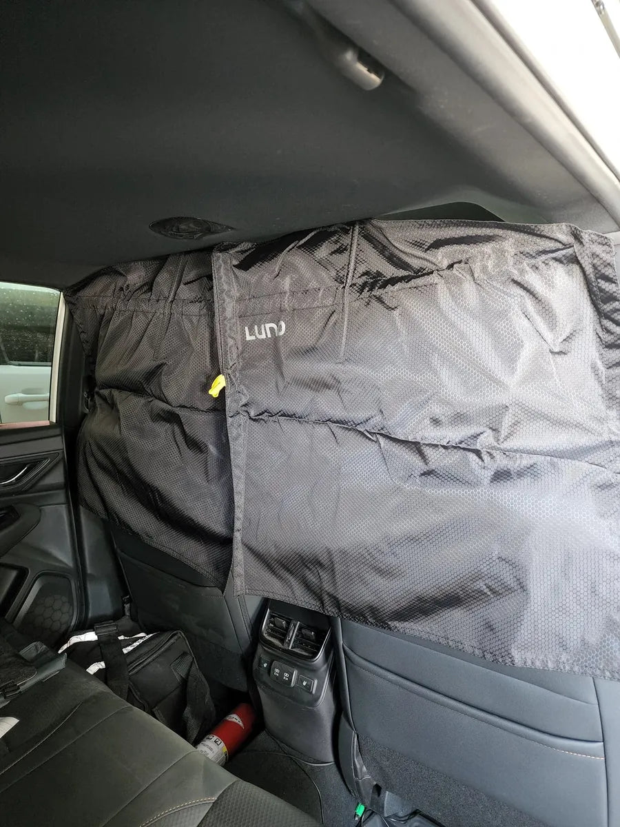 A car's backseat area with a black cover draped over the headrests, blocking the view through the rear side windows. There are various items on the seat, including a fire extinguisher and a bag. The cover has the brand name