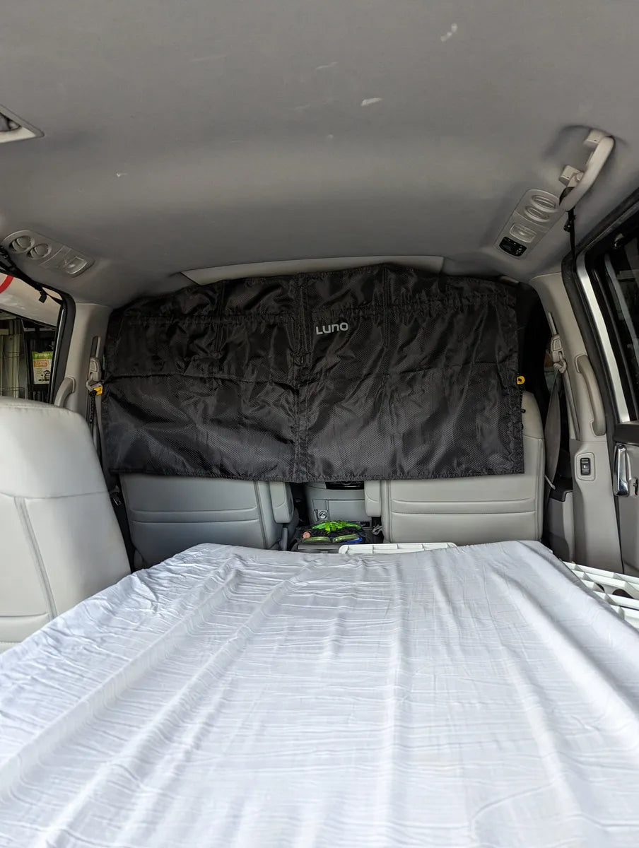 Interior view of a van with a privacy curtain installed behind the front seats. The back area includes a neatly made bed with white sheets, indicating a setup for camping or sleeping in the vehicle. The curtain has the brand name