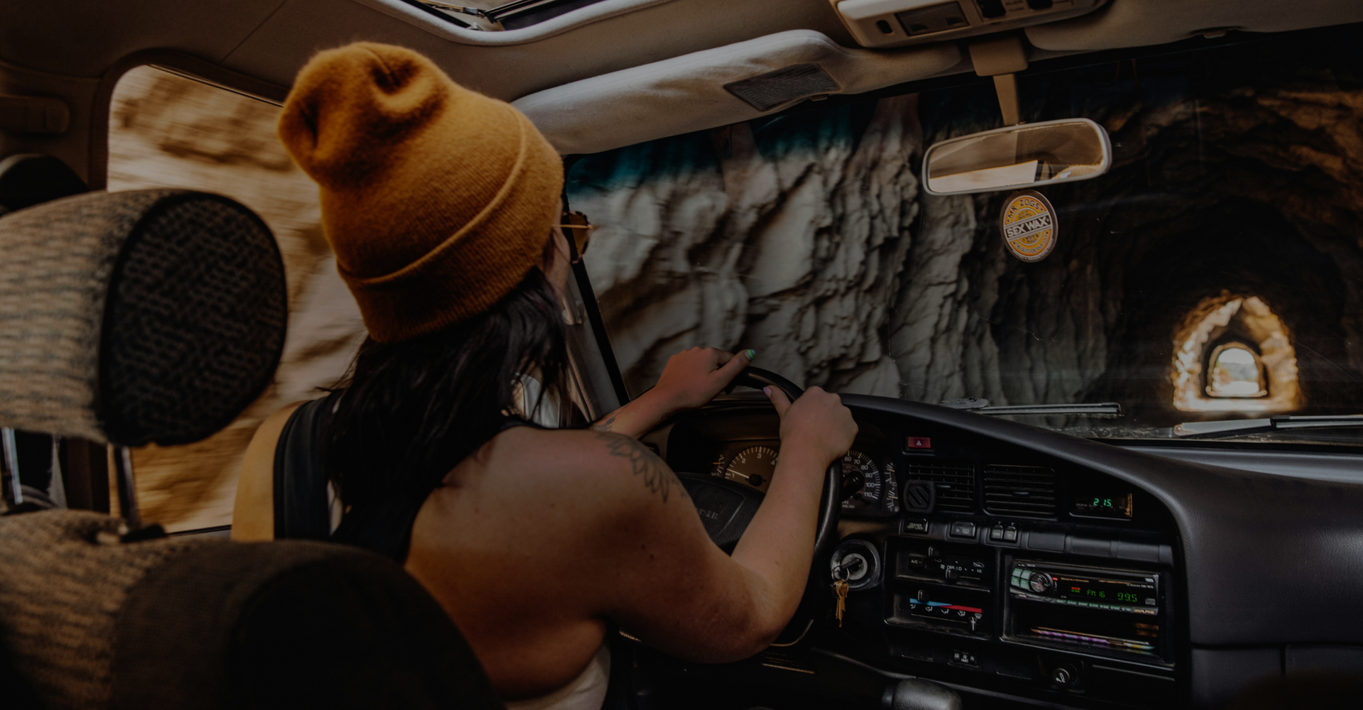 A person wearing a yellow beanie and sunglasses is driving a vehicle through a tunnel. Sunlight is visible at the end of the tunnel, illuminating the rocky interior. The headrest of the passenger seat and part of the dashboard are also visible.