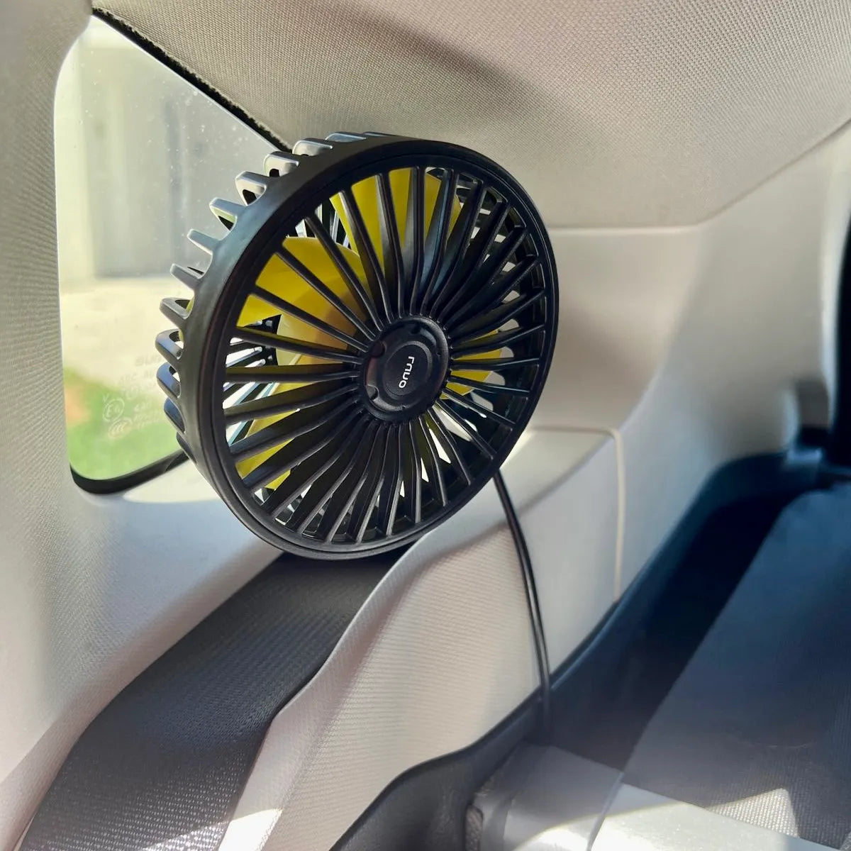 A black, portable mini fan is attached to the side window of a vehicle interior. It is plugged in with a USB cable, providing airflow within the car. The fan has a compact and circular design suitable for travel or in-car use.