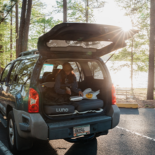An individual is sitting in the back of a blue SUV with the trunk open. The trunk is equipped with a Luno car mattress, and the person appears to be organizing or preparing something. The scene is set in a park area by a lake with sunlight breaking through the trees.
