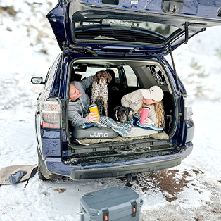 A couple relaxes in the back of an open SUV parked in a snowy area, each holding a drink. Two dogs are with them, one sitting between them and the other lying down. A gray cooler is on the ground nearby. The scene looks cozy despite the cold surroundings.