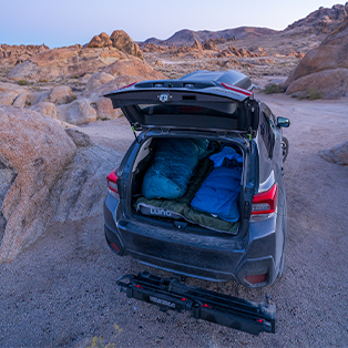 An SUV parked in a rocky desert landscape with its rear hatch open. The trunk area is set up for sleeping, featuring two sleeping bags laid out side by side. The surrounding area showcases rugged terrain and distant mountain ranges under a calm sky.