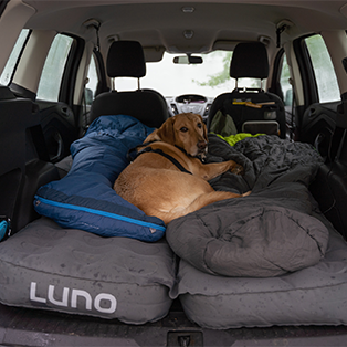 A dog lies on an inflated Luno mattress placed in the back of an SUV, surrounded by a sleeping bag and blankets. The open vehicle hatch reveals a rainy backdrop, indicating a cozy setup for a camping or road trip.