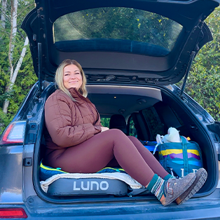 A woman sits in the back of an open hatchback car, resting on a Luno mattress. She is dressed in a brown jacket and maroon leggings, smiling contentedly. Beside her are colorful bags and the background features green foliage.