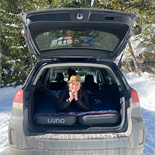 A person wearing a yellow hat and a black jacket is lying in the back of an SUV with the trunk open. They are smiling and resting their head on their hands. The vehicle is parked in a snowy area with trees in the background. The SUV has a LUNO mattress inside.