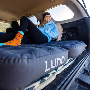 A person wearing a blue hoodie and colorful socks is lying comfortably on a mattress inside a vehicle with an open hatch. The mattress has "LUNO" written on it. The scene appears relaxed and cheerful.