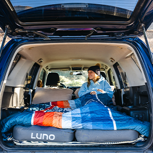A person wearing a beanie and blue jacket is sitting inside the back of a parked vehicle, which has its rear hatch open. They are sitting on a cushioned bed setup with a colorful quilt. The scene shows a cozy camping environment inside the vehicle.