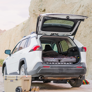 A white SUV is parked with its rear hatch open, revealing a bed setup inside. The vehicle is situated in a rocky, desert landscape. Beside the car, there is a beige cooler on the ground, and various colorful items are partially visible.