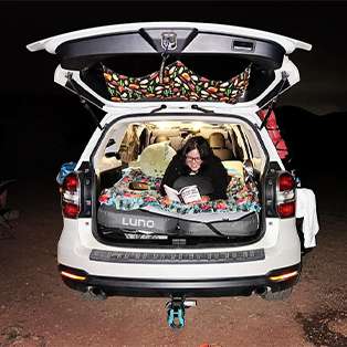 A person is reading a book while lying in the back of a white SUV, which has its trunk open. The car is set up for camping with a colorful blanket, pillows, and a hanging organizer. The scene is illuminated by interior car lights against a dark night background.