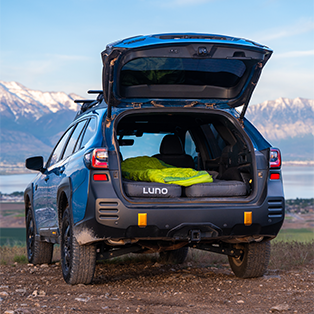 A blue SUV with its rear hatch open, revealing a neatly arranged camping setup inside. There is a green sleeping bag laid out on an elevated sleeping platform in the cargo area. The vehicle is parked in a scenic outdoor location with mountains and a lake in the background.
