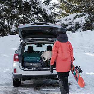 A person in a red jacket and black helmet carries a snowboard while walking towards a car with an open trunk. The car is parked in a snowy area surrounded by trees, and the trunk contains snowboarding gear.