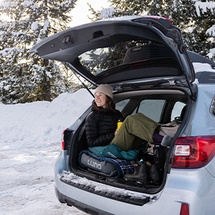 A person wearing a beanie and warm clothes sits in the back of an open SUV. The back is converted into a cozy sleeping area with blankets and pillows. Snowy trees and a snow-covered landscape are visible in the background, indicating a winter setting.