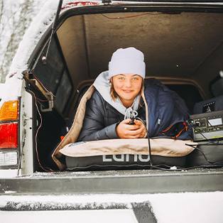 A person wearing a white beanie and dark jacket is lying in the back of a vehicle. They are smiling and holding a device. Equipment is visible beside them, and the area outside the vehicle appears to be snowy.