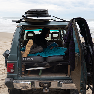 A person lies on a makeshift bed inside the back of a parked SUV at the beach, reading a book. Surfboards are strapped to the roof and another is inside the vehicle. The vehicle's back door is open, revealing a blue sleeping pad. The ocean is visible in the background.