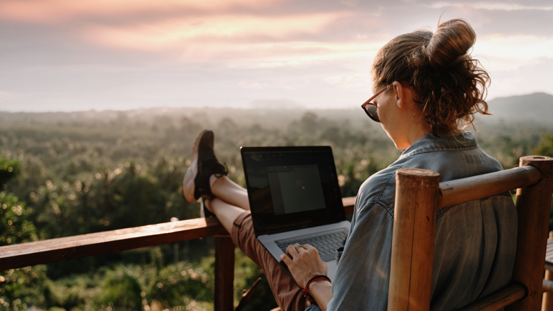 A person wearing sunglasses and a bun hairstyle sits on a wooden chair outdoors, using a laptop with their feet propped up on a railing. A lush green landscape and a serene sunset sky are in the background.