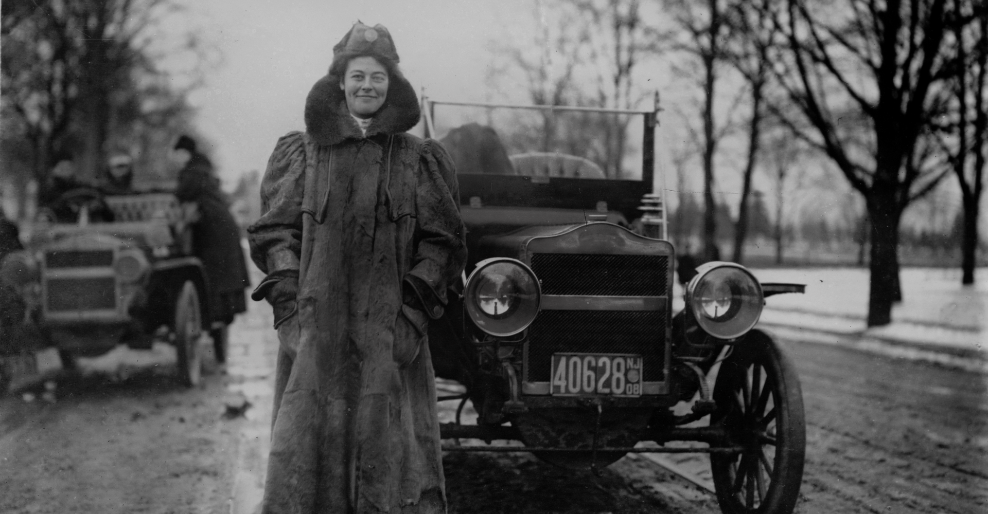 A person wearing a large coat and a hat stands in front of an early 20th-century car on a snowy road. The car has a visible license plate number "40628." Another individual and a car can be seen in the background to the left. Trees line the road.