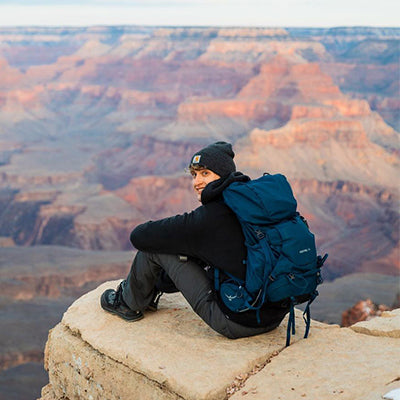 A person wearing hiking gear and a beanie sits on the edge of a rocky cliff, overlooking the expansive landscape of the Grand Canyon. They have a blue backpack and gaze into the distance, capturing a moment of solitude and contemplation.