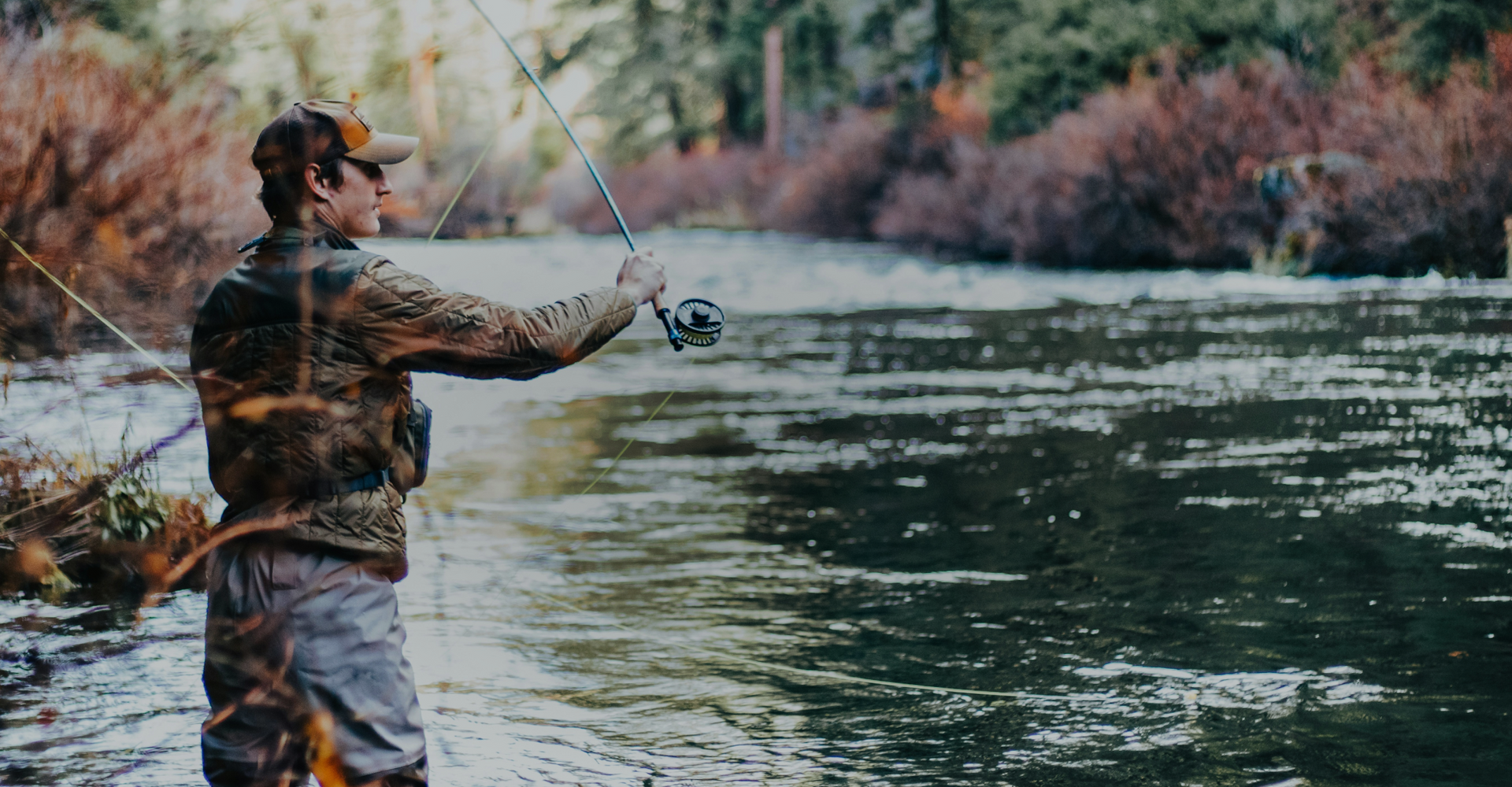 A person wearing a cap casts a fishing line into a flowing river surrounded by trees with autumn foliage. The individual stands at the riverbank, focusing on their fishing. The background features a blend of green and brown tones, typical of a forest setting.