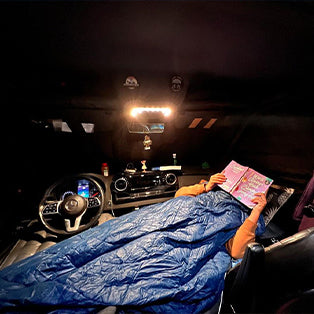 A person is lying in the front passenger seat of a car, covered with a blue sleeping bag, and reading a book. The car's dashboard and steering wheel are visible, and the interior light is on, illuminating the area. The person appears to be relaxed and comfortable.