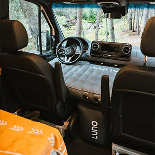 Interior of a van converted for camping, featuring an inflatable mattress positioned across the front seats. The dashboard and steering wheel are visible through the windshield. Two upholstered front seats flank the mattress, and a blanket is partially visible.